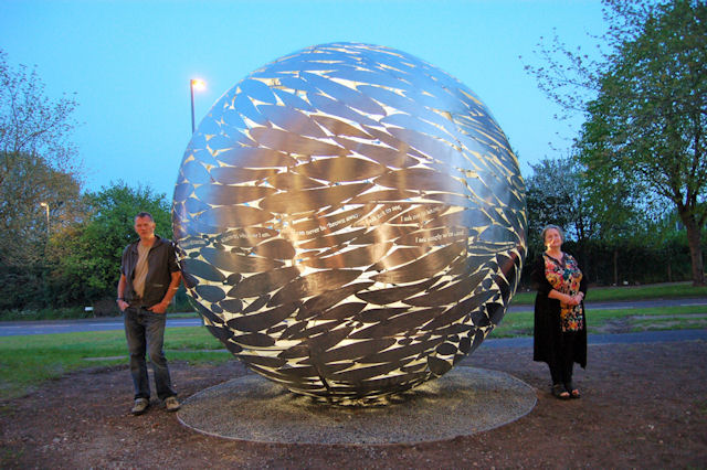 Two people stand either side of a giant metallic lattice ball in the evening