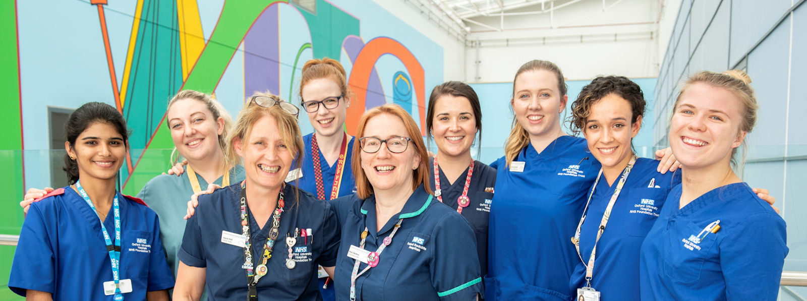 Nine smiling women in nurses' uniforms, large glass-roofed atrium behind