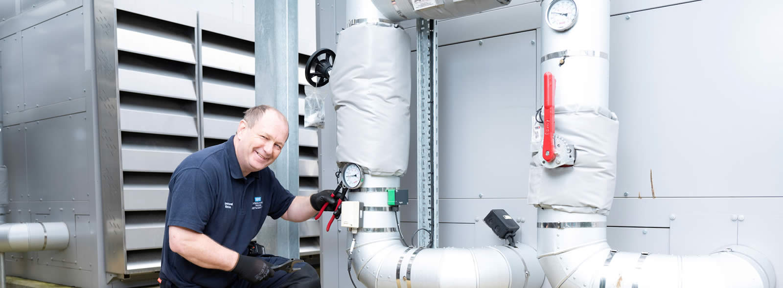 Kneeling man in work shirt and gloves adjusts gauge on a large lagged pipe