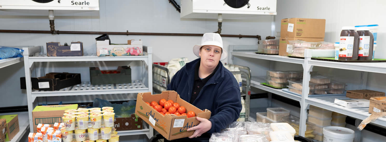 Woman in fleece and brimmed hat holding a crate of oranges in a store room