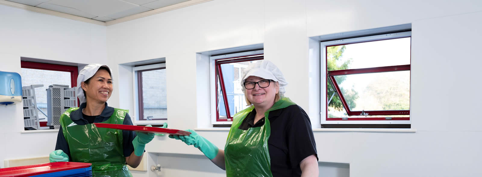 Two smiling women in plastic aprons, gloves and hats in a room with open windows, passing a tray