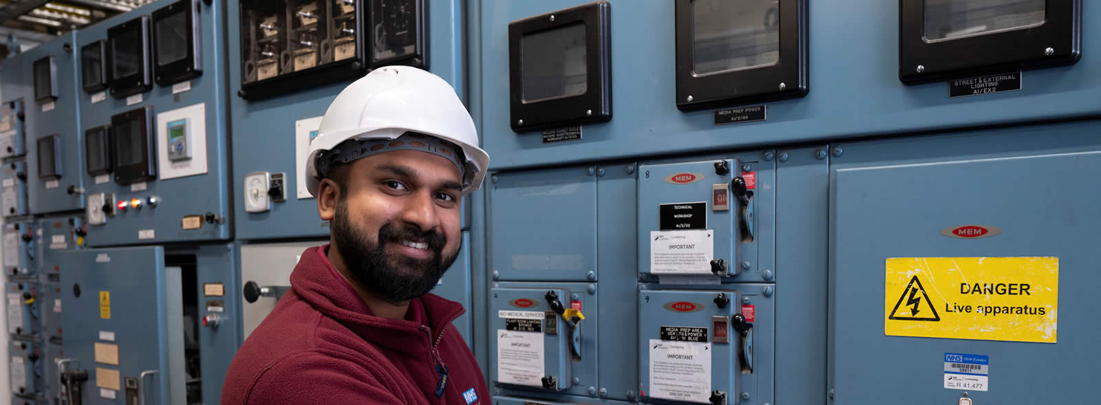 Smiling man of colour with beard and hard hat standing by wall of electical equipment displays