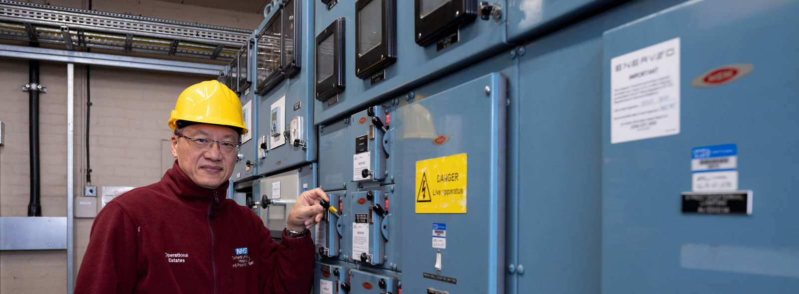 Man in hard hat and fleece stands by a bank of electrical equipment